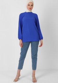 Maglietta azzurra a maniche lunghe con scollo alto, abbinata a jeans attillati azzurri chiari. La maglietta presenta maniche arricciate e una texture di tessuto liscia.