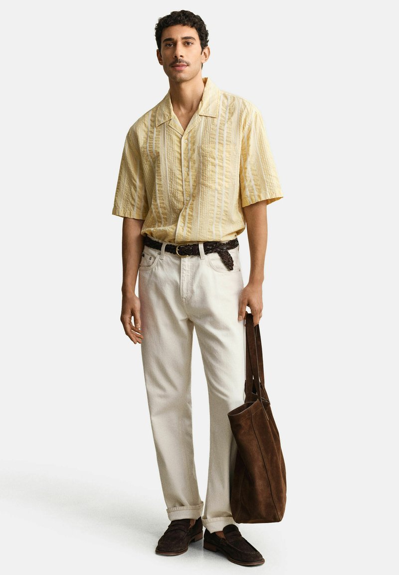 Mann trägt ein gelbes gestreiftes Kurzarmhemd, weiße Hose, braunen Gürtel und Schuhe, hält eine große braune Wildledertasche.