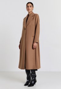 Cappotto lungo in miscela di lana colore tan, con rever a vista, bottone singolo e tasche laterali, abbinato a stivali neri lucidi con tacco.