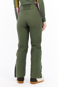 Person wearing olive green snow pants with side zippers over white and brown boots, standing against a white background.