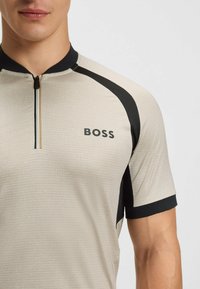 Lys beige kortærmet skjorte med sorte accenter. Har en halv lynlås og "BOSS" logoen på brystet. Struktureret stof med et subtilt mønster.