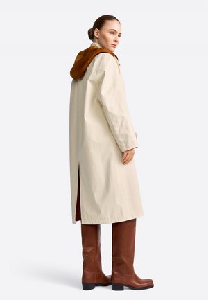 Femeie purtând un palton lung bej cu glugă maro și cizme maro din piele până la genunchi, privindu-și peste umăr.