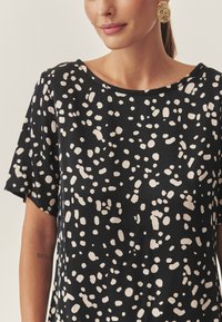 Fekete felsőrész laza szabással, rövid ujjakkal és különböző bézs formákból álló absztrakt mintával. Sima anyagból készült, kerek nyakkivágással.