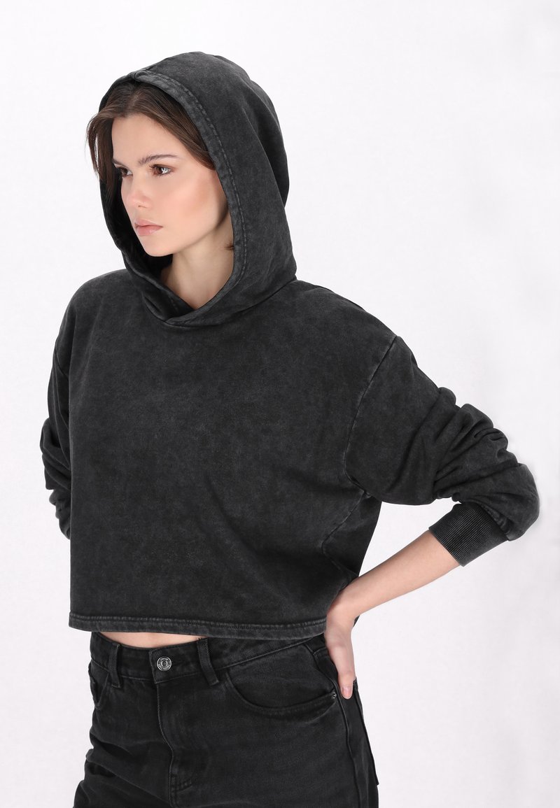 Rövidített fekete kapucnis sweatshirt, elhalványult textúrával és bordázott mandzsettával. Magas derekú fekete farmernadrággal viselve.