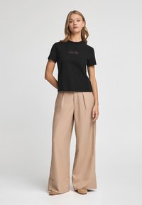 Camiseta de algodón negra de manga corta y ajuste ceñido con un logo en cursiva, combinada con pantalones beige de pierna ancha hechos de una tela suave.