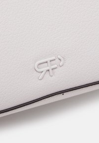 Biała teksturowana skórzana powierzchnia z wypukłym, splątanym logo "RF" w białym kolorze, umieszczonym blisko ciemnej krawędzi szwu.
