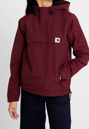 Persoon draagt een bordeauxrode capuchonjas met een voorvak en een Carhartt-logo, gecombineerd met donkerblauwe jeans.