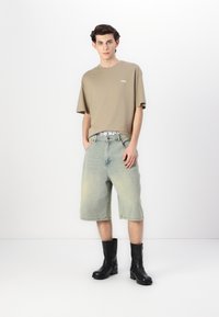 Tricou oversized khaki cu logo alb, asortat cu pantaloni scurți din denim bleu deschis. Modelul poartă ghete negre cu detalii din cataramă.