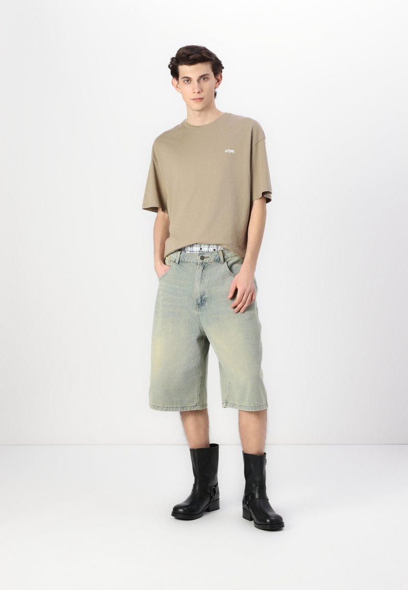 Tricou oversized khaki cu logo alb, asortat cu pantaloni scurți din denim bleu deschis. Modelul poartă ghete negre cu detalii din cataramă.
