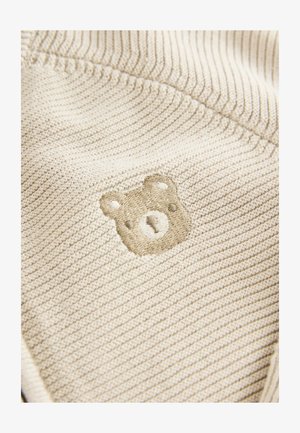 Bézs kötött anyag, bordázott textúrával; finom világosbarna medve embléma hímzett a felületén.