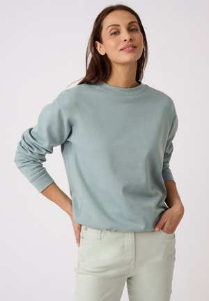 Donna con lunghi capelli castani che indossa una felpa azzurro chiaro e pantaloni chiari, in piedi con le mani nelle tasche davanti a uno sfondo uniforme.