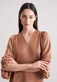 Brun V-halset bluse med puffede ærmer og samlede manchetter, lavet af et glat stof. Har et enkelt design uden bemærkelsesværdige mønstre.