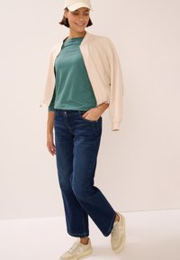 Light beige bomber jacket, teal striped shirt, dark blue jeans, and beige sneakers with white accents. Model poses with a relaxed stance.