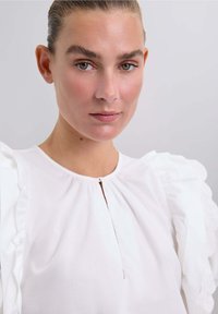 Weiße Bluse mit Puffärmeln, runder Ausschnitt und einem Schlitzdetail vorne. Weicher Stoff mit subtiler Textur.