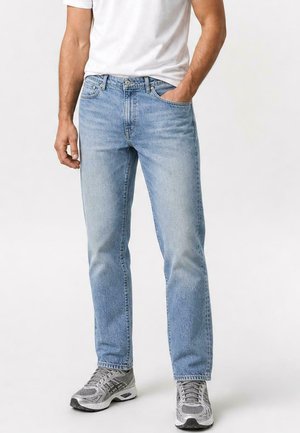 Man draagt lichtblauwe straight-fit jeans, wit T-shirt en grijze hardloopschoenen, staat met één hand in de zak.