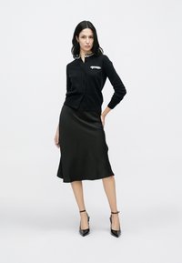 Black cardigan with sequined collar detail, paired with a black satin slip skirt. Model wears black pointed-toe heels and has long dark hair.