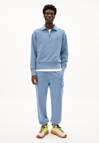 Lichtblauwe sweatshirt met rits en een iets cropped pasvorm, gecombineerd met bijpassende cargo sweatpants. Witte T-shirt eronder en heldere sneakers.
