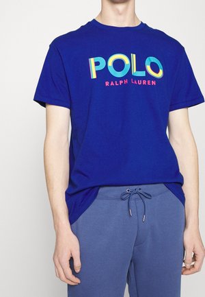 Mann trägt ein royalblaues Polo Ralph Lauren T-Shirt mit buntem Logo und blaue Sweatpants mit Kordelzug, steht vor einem einfarbigen Hintergrund.