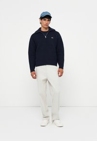 Marineblauwe gebreide trui met kwart-rits en geribbelde details, gecombineerd met lichtgrijze broeken en witte sneakers. Een denim pet maakt de outfit compleet.
