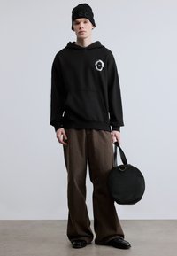 Μαύρο φούτερ με κουκούλα και λεπτομέρεια λογότυπου, χαλαρή εφαρμογή, τσέπη καγκουρό· καφέ παντελόνι φαρδιάς γραμμής· μαύρη στρογγυλή τσάντα· μαύρο ριμπ σκουφάκι.