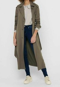 Kobieta ubrana w oliwkowy płaszcz trenchowy, koszulę w kratę, ciemnoniebieskie obcisłe dżinsy i beżowe sneakersy, stojąca na tle jednolitej kolorystyki.