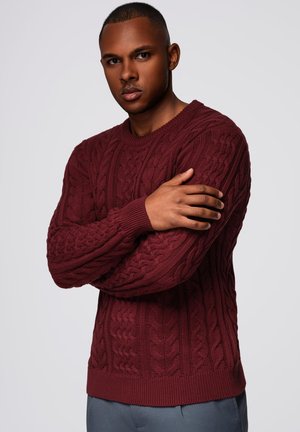 Bordeauxroter Kabelstrickpullover mit rundem Ausschnitt, strukturiertem Muster und gerippten Ärmeln und Saum, getragen über grauen Hosen.