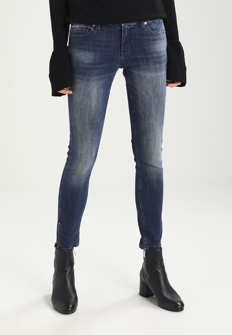 Jeans ajustados de mezclilla de color azul oscuro, con un desgaste sutil y cremalleras en los tobillos, combinados con botas de tacón negro hasta el tobillo.