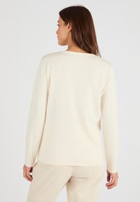 Jersey de punto color crema de manga larga con escote redondeado, textura suave y corte relajado. Llevado con pantalones de canalé de color claro.