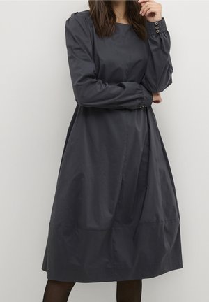 Kvinde iført en langærmet, mørkegrå midi-kjole med knapmanchetter, stående mod en ensfarvet lys baggrund, med hånden nær ansigtet.