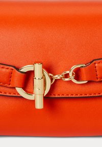 Narancssárga bőrtáska arany színű, kör alakú zárással, vékony fekete pánttal, és textúrált felülettel, amelyen látható varrási részletek találhatók.