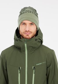 Grüne wasserdichte Jacke mit hohem Kragen, mit Reißverschlusstaschen und Belüftungsöffnungen, kombiniert mit einer hellgrünen Strickmütze mit Logodetail.
