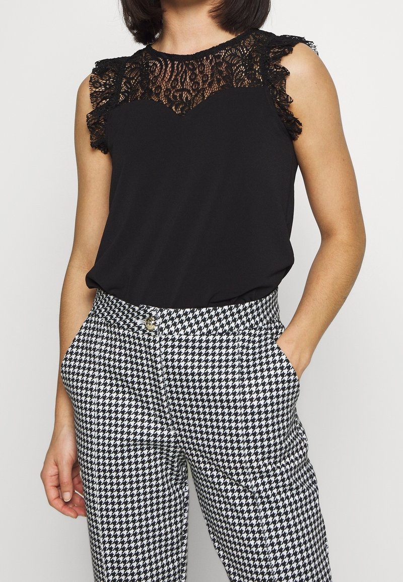 Femme portant un haut en dentelle noir sans manches associé à un pantalon taille haute à motif pied-de-poule noir et blanc.