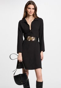 Abito nero con colletto, con bottoni sul davanti, stretto in vita da una cintura con accenti dorati lucidi, abbinato a una piccola borsa nera.