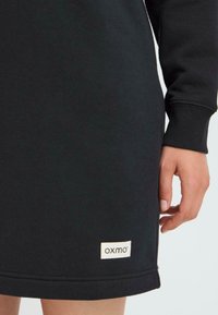 Musta college-mekko pitkilla hihoilla, ribbatut hihansuut ja pyöristetty helma. Siinä on pieni valkoinen etiketti "oxmo" lähellä helmaa.
