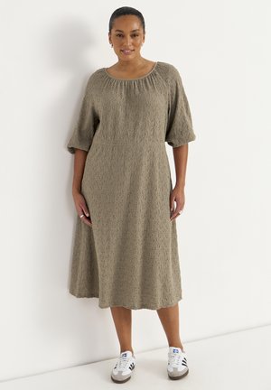 Kvinde står iført en tekstureret, løstsiddende, knælængde olivengrøn kjole med halvlange ærmer og hvide Adidas sneakers.