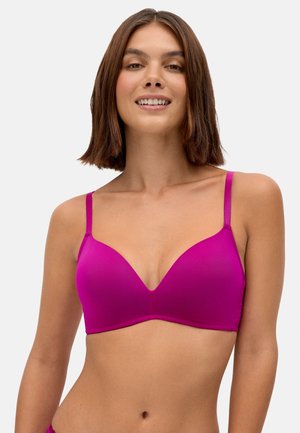 Mujer con cabello castaño hasta los hombros sonriendo, vestida con un sujetador rosa brillante con tirantes ajustables, sobre un fondo claro y sencillo.