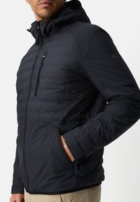 Zwarte jas met capuchon, voorzien van een doorgestikte bovenkant, een gladde onderkant, ritssluitingen in de zakken en elastische manchetten met een strakke pasvorm.