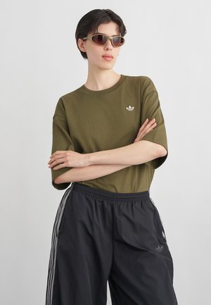 Person iført olivengrøn Adidas t-shirt og sorte Adidas træningsbukser med krydsede arme og ovale solbriller, foran en ensfarvet baggrund.
