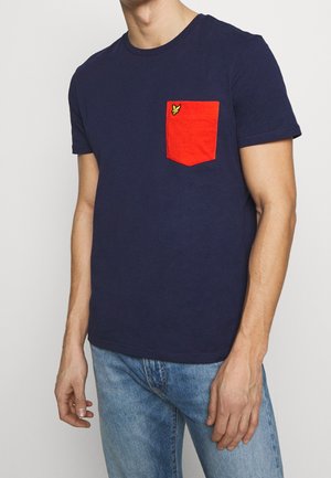 Mann trägt ein marineblaues kurzärmeliges T-Shirt mit einer leuchtend roten Brusttasche, die ein kleines gelbes Logo zeigt, kombiniert mit hellblauen Jeans.