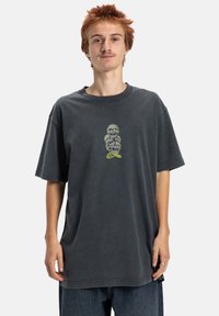 Tmavě šedé oversized tričko vyrobené z bavlny, s grafikou postavy v zelené a černé barvě na hrudi, s krátkými rukávy.