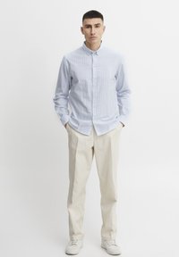 Camisa de botón a rayas azul claro hecha de algodón, combinada con pantalones beige y zapatillas blancas. Cuenta con un cuello clásico y cierre de botón.