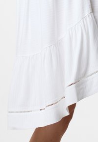 Weißes Kleid mit gestuftem Saum, das eine subtile gestreifte Textur und zarte Ausschnittdetails am unteren Rand aufweist.
