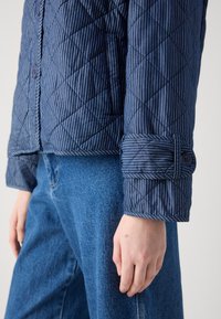 Gequilted marineblauwe jas met witte strepen, voorzien van een knoopsluiting aan de manchetten en een lichtgewicht, gestructureerde stof. Gecombineerd met blauwe jeans.