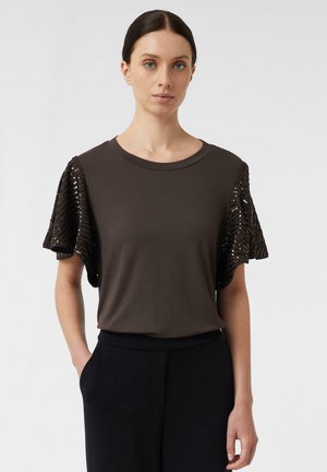 Kobieta o ciemnych włosach spiętych do tyłu, ubrana w ciemnobrązową bluzkę z krótkimi, falbaniastymi rękawami ozdobionymi cekinami oraz czarne spodnie.