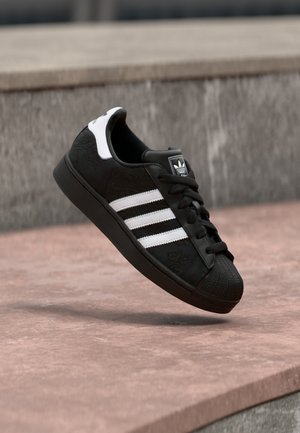 Μαύρο αθλητικό παπούτσι Adidas με λευκές ρίγες και ταμπελάκι στη φτέρνα, αιωρούμενο στον αέρα πάνω από ροζ πέτρινη επιφάνεια και γκρι τσιμεντένιο φόντο.