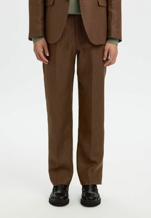 Osoba nosząca brązowe, szyte na miarę spodnie, pasującą marynarkę, widoczne zielone mankiety swetra oraz czarne skórzane mokasyny, stojąca na białym tle.