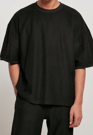 Homme portant une chemise noire côtelée oversize à manches courtes et un pantalon assorti, debout devant un fond neutre avec les mains détendues.