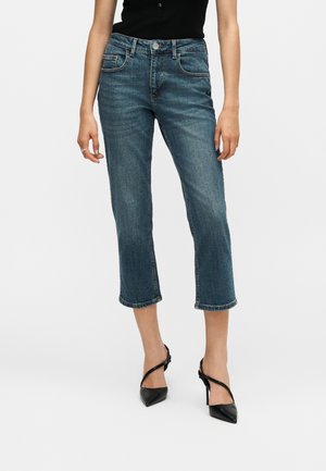 Kvinde iført afklippede blå denim-jeans, sort ærmeløs top og sorte spidse højhælede sko står mod en ensfarvet baggrund.