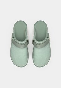 Ανοιχτό πράσινα ξύλινα παπούτσια με απαλό υλικό, διαθέτουν φαρδιά λουρί στο επάνω μέρος και ραφές σε αντίθεση κατά μήκος των άκρων. Μινιμαλιστικός σχεδιασμός.