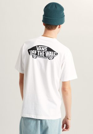 Persoon die een wit Vans "Off The Wall" t-shirt draagt, gezien van achteren, blauwe gebreide muts en lichtblauwe broek tegen een effen achtergrond.